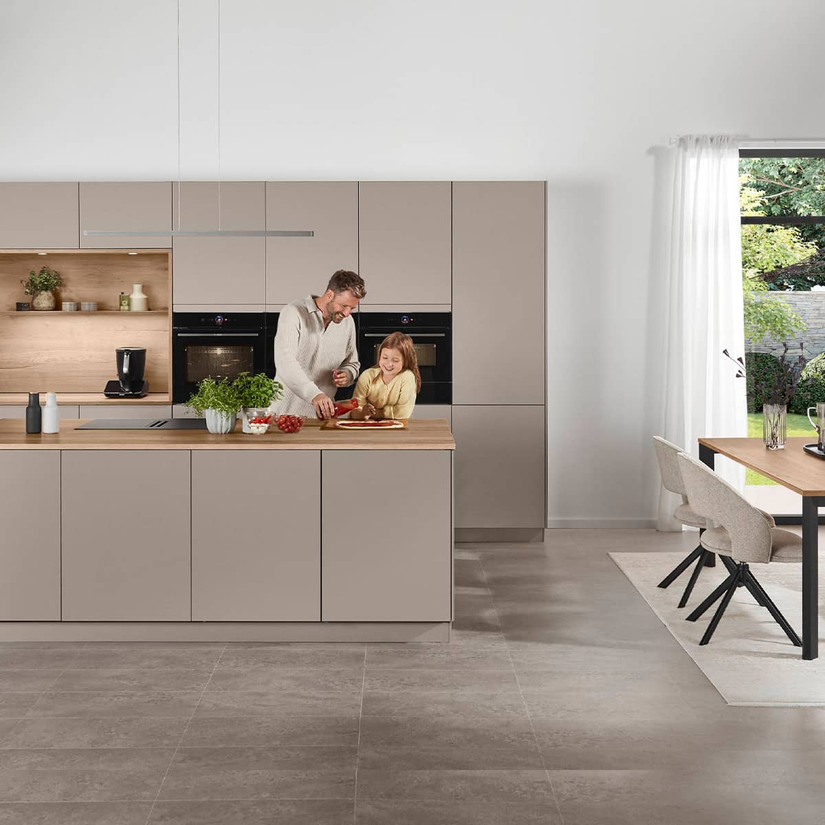 Senso Kitchen 483 with human element Minimalist taupe kitchen design with a large island, wooden breakfast bar, and integrated black ovens in a bright, open-plan home.
