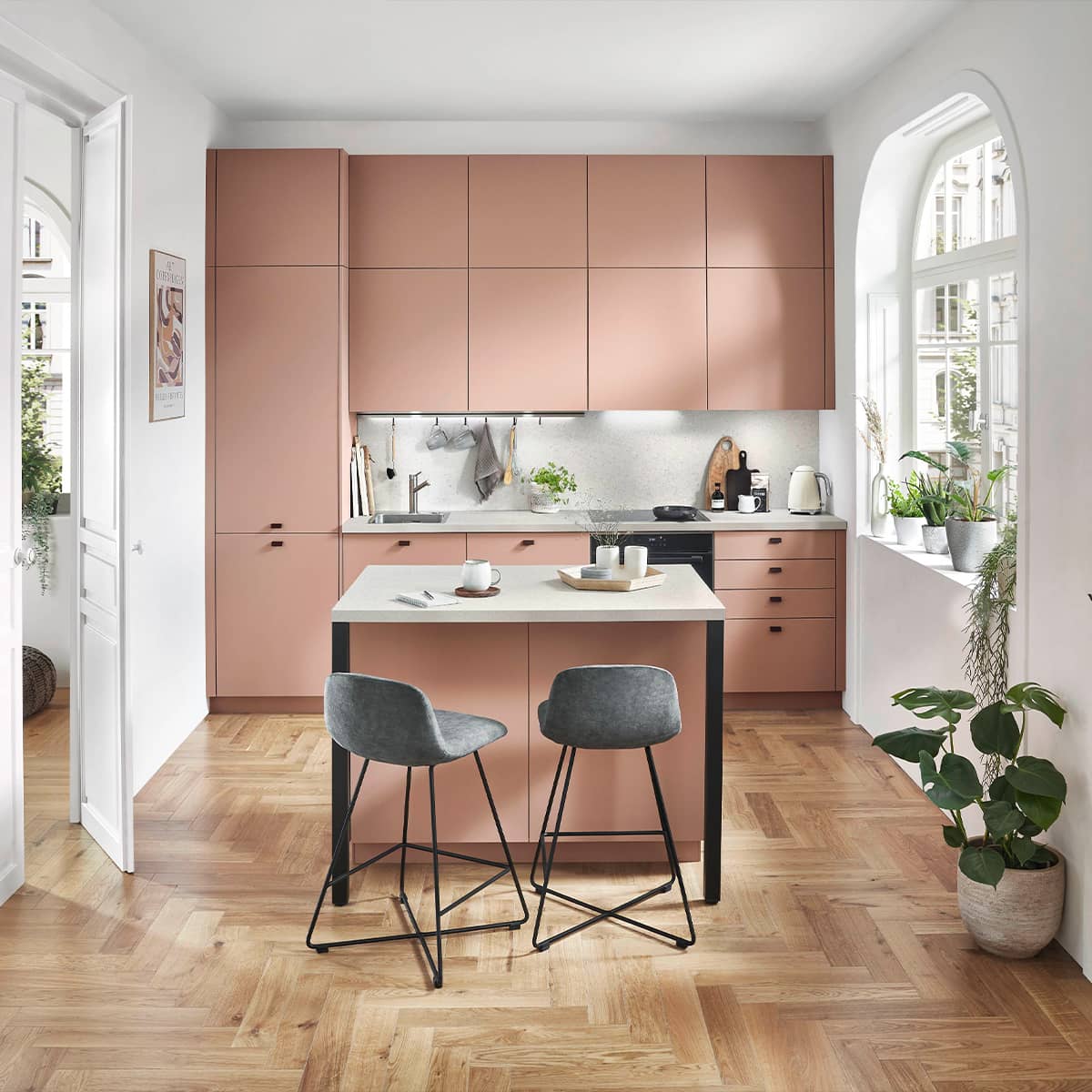 Senso Kitchen 492 - Coral Salmon Kitchen Compact salmon pink minimalist kitchen with handleless cabinets, a small matching island, and grey bar stools on a herringbone wood floor.