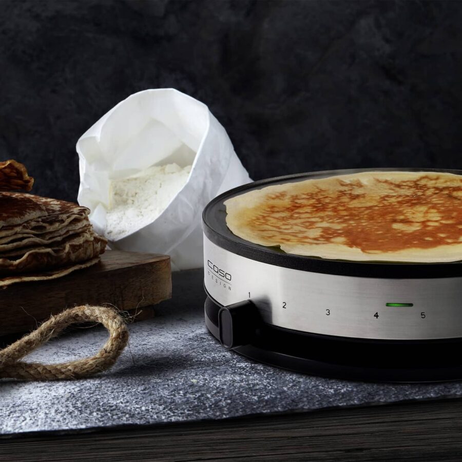 Caso Crepes Maker CM 1300 on countertop with crepe cooking, flour bag and stacked crepes in background.